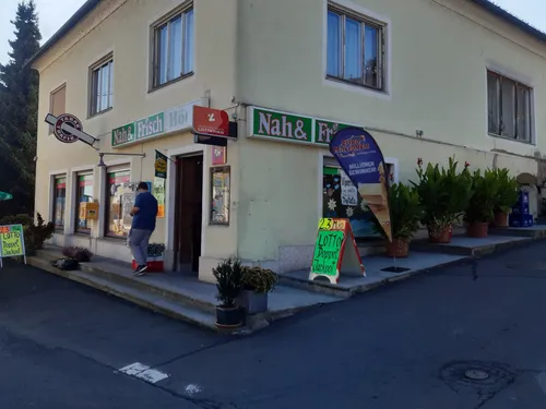 Ein Gebäude mit einem Lotterieladen, 'Nah & Frisch' und 'Lotterien' Schildern. Ein Mann steht in der Nähe des Eingangs, und ein Schild für 'Lotto Doppel Jackpot' ist draußen.