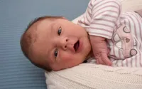 Ein Neugeborenes liegt auf einer Decke und trägt ein rosa-weiß gestreiftes Langarmshirt. Das Baby hat einen überraschten Ausdruck, die Augen weit geöffnet und der Mund leicht geöffnet.