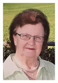 Eine Frau mit einer Brille und einer Halskette mit Kreuzanhänger steht draußen. Sie lächelt und scheint für ein Foto zu posieren. Sie trägt ein grünes Hemd und hat kurze Haare. Hinter ihr ist ein verschwommener Hintergrund mit einem Rasenfeld und einigen Pflanzen.