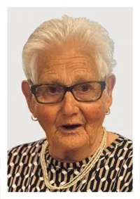 Eine ältere Frau mit weißen Haaren, die eine Brille, eine Halskette und Ohrringe trägt, posiert für ein Foto. Sie hat einen ernsten Ausdruck und trägt ein schwarz-weiß gemustertes Oberteil.