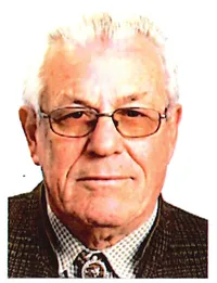 Ein älterer Mann mit weißem Haar und einem Schnurrbart, der eine Brille und eine karierte Jacke trägt, lächelt leicht in die Kamera.