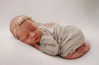Ein Babymädchen schläft auf einer weißen Oberfläche, eingewickelt in einen hellen Stoff und mit einer Blumenhaarspange.