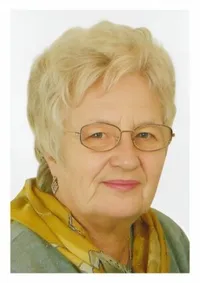 Ein Porträt einer älteren Frau mit kurzen blonden Haaren, die eine Brille und ein gelbes Tuch trägt. Sie hat einen neutralen Gesichtsausdruck und steht vor einem schlichten Hintergrund.