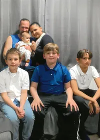 Eine Familie mit fünf Personen posiert für ein Foto. Ein Mann hält ein Baby, während eine Frau hinter ihm steht. Drei Kinder sitzen auf einer Couch, zwei in weißen Hemden, eines in Blau.