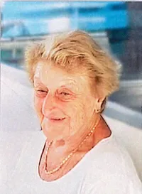 Eine ältere Frau mit blondem Haar, die eine Halskette trägt, schaut direkt in die Kamera. Der Hintergrund ist unscharf mit Anzeichen von Blau.