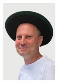 Ein Mann mit schwarzem Hut und weißem T-Shirt lächelt in die Kamera. Er hat kurze, helle Haare und steht vor einem weißen Hintergrund.