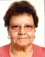 Ein Nahporträt einer älteren Frau mit kurzen lockigen Haaren, die eine Brille und ein rosa Oberteil trägt und direkt in die Kamera mit einem neutralen Gesichtsausdruck schaut.