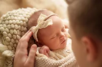Ein Nahaufnahme eines schlafenden Neugeborenen mit einer Spitzenhaarschleife, das von einem Elternteil in den Armen gehalten wird. Das Baby ist in eine beige gestrickte Decke gewickelt.