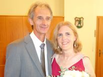 Ein Mann und eine Frau stehen eng beieinander und lächeln für ein Foto. Der Mann trägt einen grauen Anzug und eine Krawatte, während die Frau ein rotes Kleid trägt und einen Blumenstrauß hält. Hinter ihnen befindet sich eine gelbe Wand mit einem dekorativen Emblem.