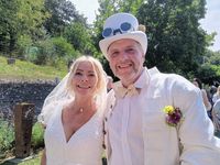 Ein Mann und eine Frau in weißen Kleidern, die eng beieinander stehen und lächeln, mit einem grünen Hintergrund aus Bäumen und einem Zaun.