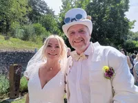 Ein Mann und eine Frau in weißen Kleidern, die eng beieinander stehen und lächeln, mit einem grünen Hintergrund aus Bäumen und einem Zaun.