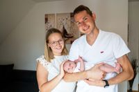 Ein lächelndes Paar mit einem Neugeborenen, der Vater trägt ein weißes Levi's-Shirt und die Mutter ein weißes Oberteil, stehen in einem Raum mit weißen Wänden und einer Couch.