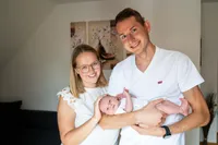 Ein lächelndes Paar mit einem Neugeborenen, der Vater trägt ein weißes Levi's-Shirt und die Mutter ein weißes Oberteil, stehen in einem Raum mit weißen Wänden und einer Couch.