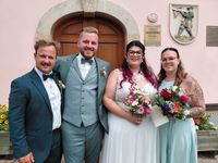 Eine Braut mit rosa Haaren und einem Hochzeitskleid steht zwischen zwei Trauzeugen und hält einen bunten Blumenstrauß. Sie posieren vor einem rosa Gebäude mit einer Holztür und dekorativen Plaketten.