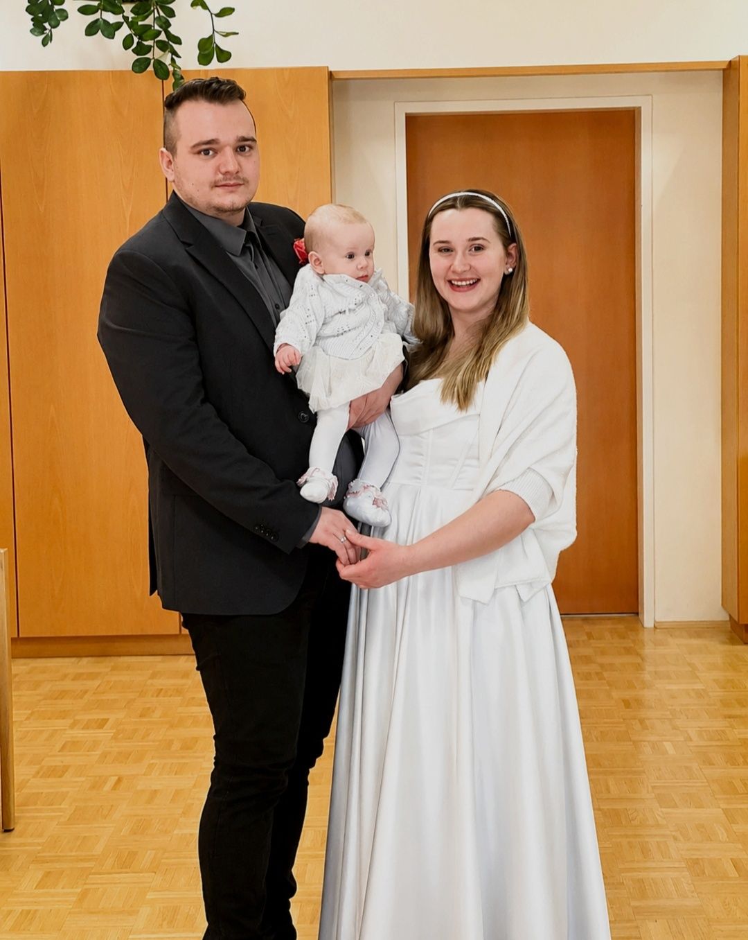 Ein lächelndes Paar hält ein Baby, gekleidet in Weiß, im Innenraum mit Holzwänden und Holzboden.