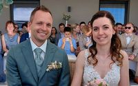 Ein lächelndes Paar bei einer Hochzeit, mit Gästen im Hintergrund. Ein Junge macht ein Foto mit seinem Handy.