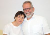 Ein älterer Mann mit grauem Haar und Bart, der eine Brille und ein weißes Hemd trägt, lächelt neben einer Frau mit kurzen Haaren, die ebenfalls lächelt und ein weißes Hemd trägt. Sie stehen vor einer weißen Wand.