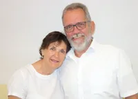 Ein älterer Mann mit grauem Haar und Bart, der eine Brille und ein weißes Hemd trägt, lächelt neben einer Frau mit kurzen Haaren, die ebenfalls lächelt und ein weißes Hemd trägt. Sie stehen vor einer weißen Wand.