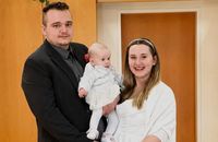 Ein Mann, eine Frau und ein Baby posieren für ein Foto. Der Mann ist formell gekleidet, und die Frau hat eine Stirnband. Das Baby trägt Weiß.
