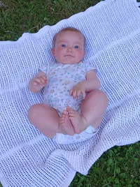 Ein Baby liegt auf einer weißen Decke mit einem Blumenhemd und schaut direkt in die Kamera. Die Decke hat ein gepunktetes Muster.
