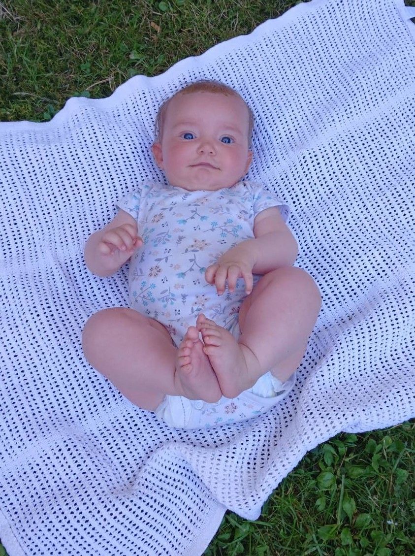 Ein Baby liegt auf einer weißen Decke mit einem Blumenhemd und schaut direkt in die Kamera. Die Decke hat ein gepunktetes Muster.