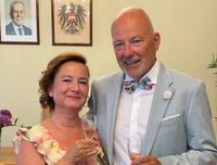 Ein Mann und eine Frau stehen nah beieinander und lächeln für die Kamera. Sie halten Weingläser in den Händen. Der Mann trägt eine Fliege und die Frau einen Halskette. Hinter ihnen hängen zwei Bilderrahmen an der Wand.