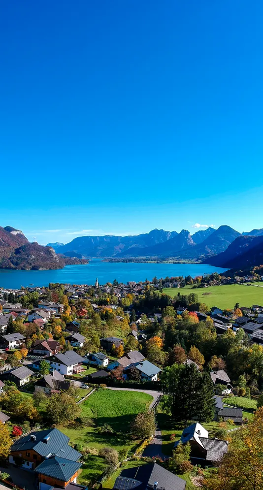 Abschnittsbild: Luftaufnahme eines Dorfes mit einem See und Bergen im Hintergrund. Das Dorf hat viele Häuser, einige mit Solarpanelen auf den Dächern. Der Himmel ist klar und blau.