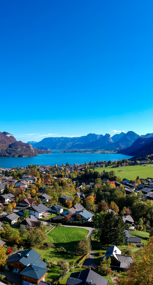  Abschnitt Bild: Luftaufnahme eines Dorfes mit einem See und Bergen im Hintergrund. Das Dorf hat viele Häuser, einige mit Solarpanelen auf den Dächern. Der Himmel ist klar und blau.
