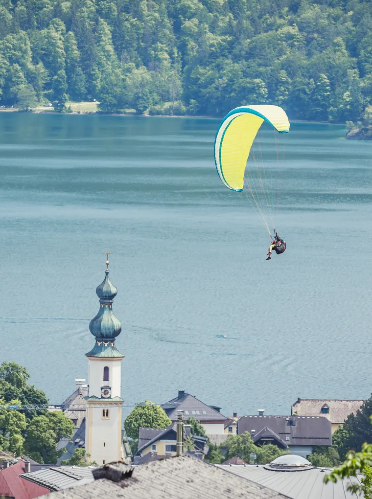 Abschnittsbild: Eine Person paraglit über einem ruhigen See mit einem Berg im Hintergrund. Ein Kirchturm ist im Vordergrund zu sehen.