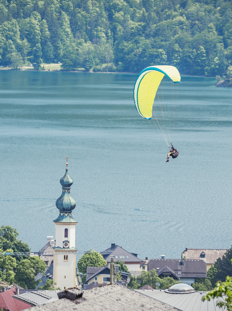  Abschnitt Bild: Eine Person paraglit über einem ruhigen See mit einem Berg im Hintergrund. Ein Kirchturm ist im Vordergrund zu sehen.