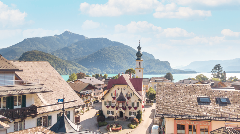  Abschnitt Bild: Luftaufnahme einer Stadt mit traditioneller Architektur. Eine Kirche mit einem Turm ist im Zentrum zu sehen. Berge und ein See sind im Hintergrund.