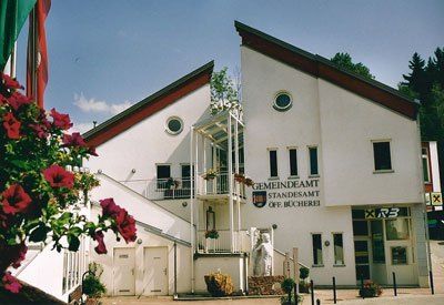 Ein weißes Gebäude mit rotem Rand hat den Text 'GEMEINDEAMT STANDESAMT DER BUCHERE' über dem Eingang geschrieben. Das Gebäude hat mehrere Fenster, eine Statue im Erdgeschoss und Topfpflanzen draußen.