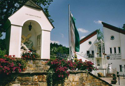 Eine kleine weiße Kapelle mit einer Jesus-Statue steht vor einem weißen Gebäude mit grünem Rand. Eine Flagge ist auf der rechten Seite. Rosa Blumen sind davor.