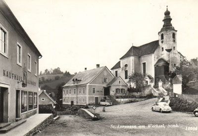 Schwarz-Weiß-Foto eines Dorfes mit einer Kirche und einem auf der Straße geparkten Auto. Die Kirche hat einen hohen Turm und ein Schild, das St. Lorenzen am Wechsel liest.