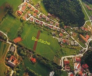 Eine Luftaufnahme eines Dorfes mit vielen Häusern und Bäumen. Ein gelber Kreis umgibt den Text Baufläche in der Mitte eines großen grünen Feldes.