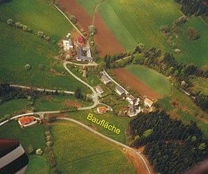 Ein Luftbild eines ländlichen Gebiets mit grünen Feldern und mehreren Gebäuden. Das Feld ist in Parzellen mit verschiedenen Nutzpflanzen unterteilt. Eine Straße verläuft durch das Feld. Der Name 'Baufläche' ist auf einer gelben Linie geschrieben.