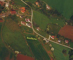 Luftaufnahme eines ländlichen Gebiets mit Häusern, Feldern und einem hervorgehobenen Bereich namens Bauflache. Grüne Felder und rote Dächer sind sichtbar.