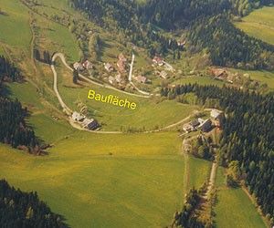Luftaufnahme eines ländlichen Gebiets namens Bauflaeche mit mehreren Gebäuden, einer Straße und üppigen grünen Feldern, umgeben von Bäumen.