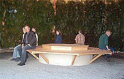 Fünf Personen sitzen auf einer Holzbank um eine große, achteckige Betonbasis in einem Außengarten.