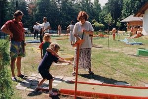 Eine Gruppe von Menschen spielt an einem sonnigen Tag im Park Minigolf. Ein Kind nimmt einen Schlag, während eine erwachsene Frau zusieht.