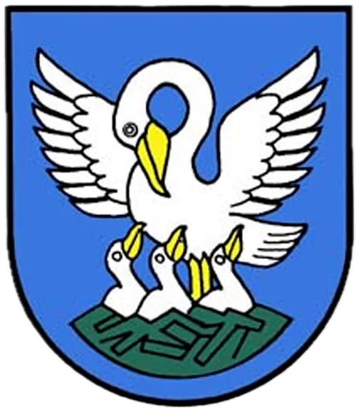 Ein weißer Schwan mit ausgebreiteten Flügeln und einem gelben Schnabel, begleitet von drei Jungschwänen, ruht auf einem grünen Banner auf einem blauen Schild.