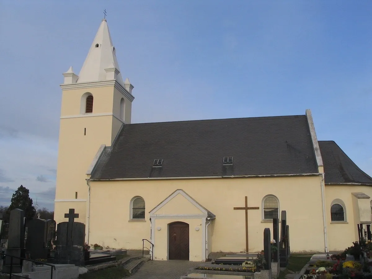 Eine Kirche mit einem Turm und einer Holztür. Ein Kreuz ist an der Wand. Gräber befinden sich an der Seite.