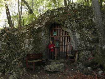 Eine Frau steht vor einer hölzernen Tür, die in eine Felswand gehauen ist, und hält ein Buch. Zwei hölzerne Bänke flankieren den Eingang.